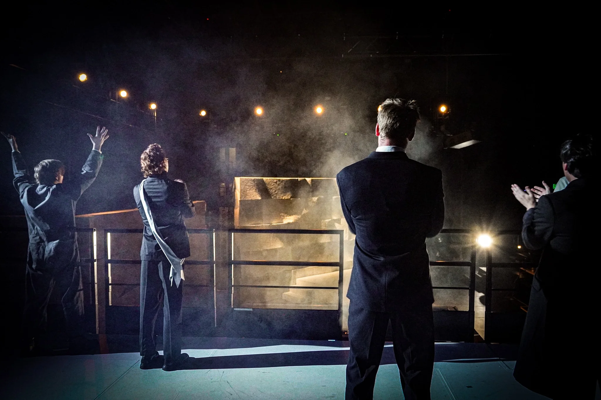Performers gather by a scaffold in a dark stage scene