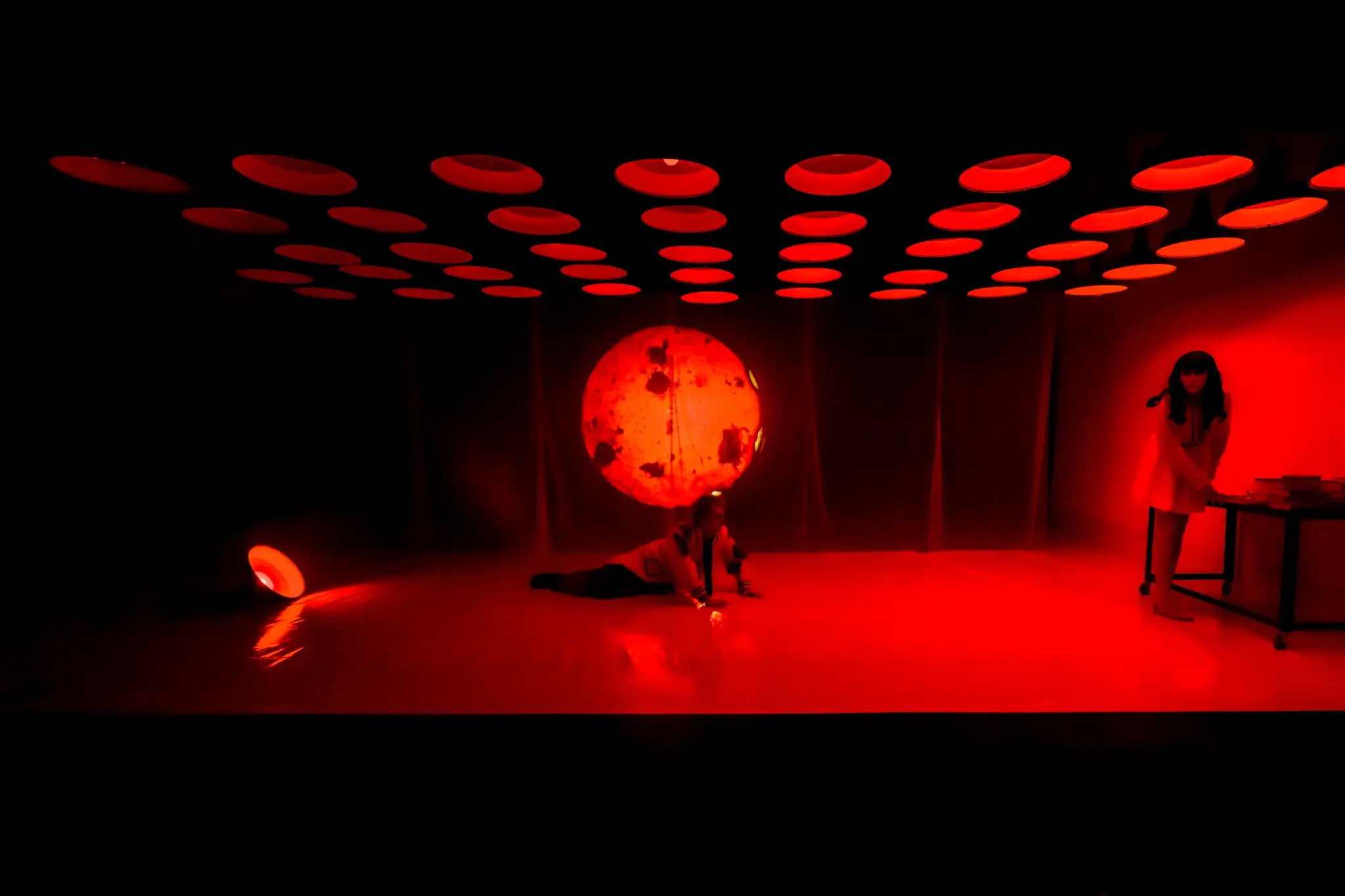 Two performers struggle beneath a red projected face