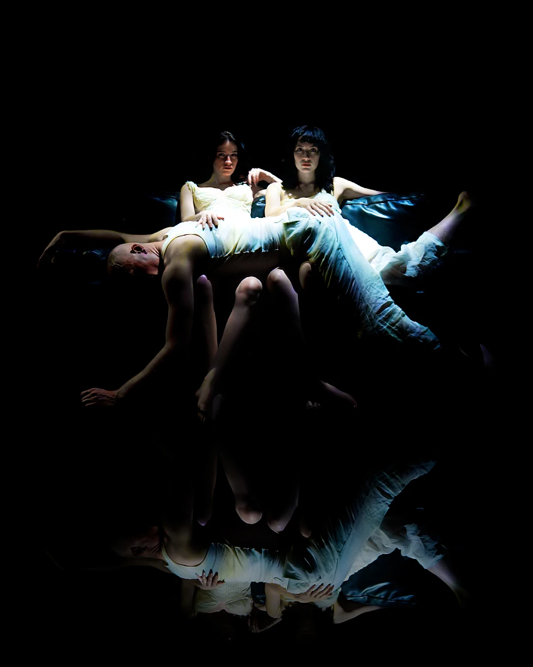 Three performers arranged across a sofa above a dark reflection