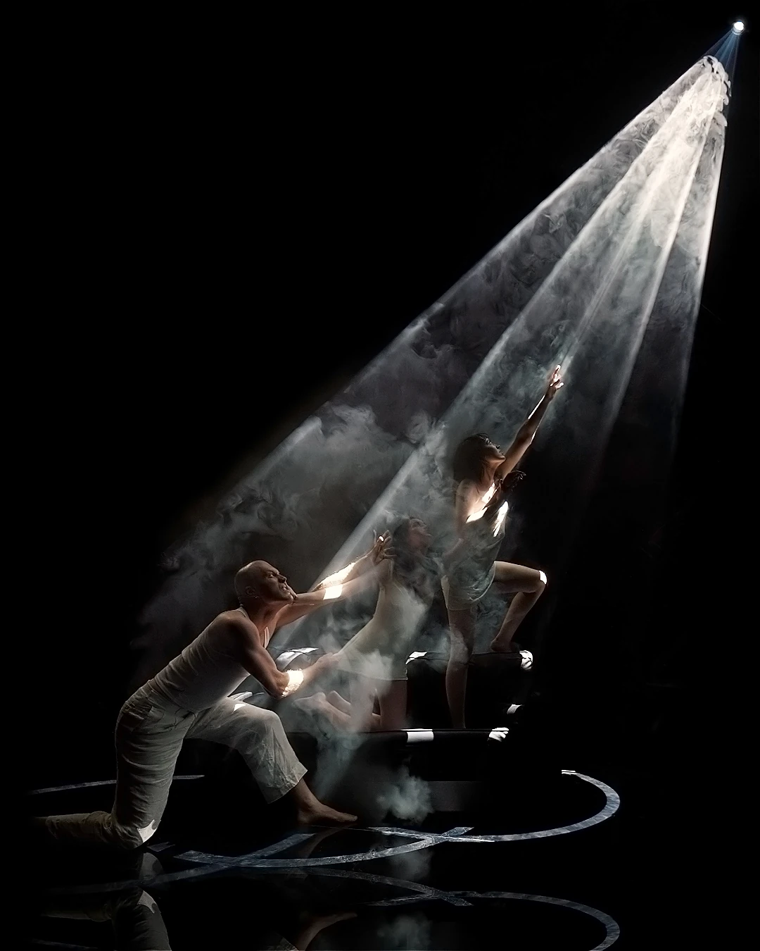 Three performers reach into a cone of light on a dark stage
